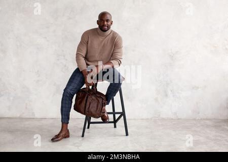 Stylish man sitting on chair and looking at camera. Confident fashion model in fashionable outfit turtleneck knitted sweater, plaid trousers, leather Stock Photo