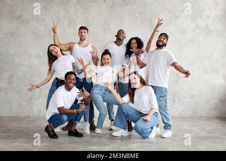 Multiethnic group of happy friends smile and raise hands up. Diverse young people standing together in white shirts and jeans on background wall in st Stock Photo