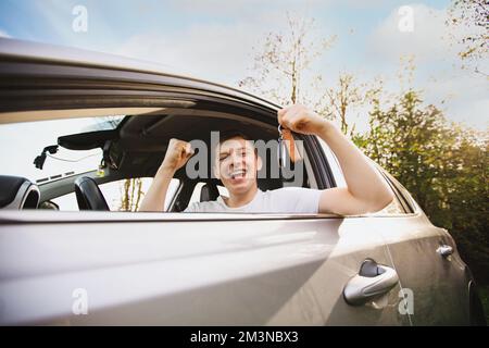 Happy Novice driver keeps fist up tight looking out of the driver window of his new car holding key from a new car. Successful young man bought a new Stock Photo