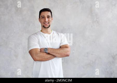 Brunette man smiles, looks at camera, crossed arms over chest on white textured wall background, natural light. Latin model in white t shirt Stock Photo