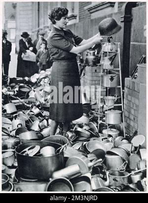 Lord Beaverbrook the Minister for Aircraft Production appealed for objects wholly or partly made of aluminium for the manufacture of aeroplanes. Photograph of a woman from the from the Women's Voluntary Service outside their town hall, surrounded by kitchen utensils including a German steel helmet from World War One. Stock Photo