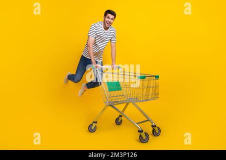 Full length photo of impressed funky man wear white t-shirt jumping high walking supermarket basket isolated yellow color background Stock Photo