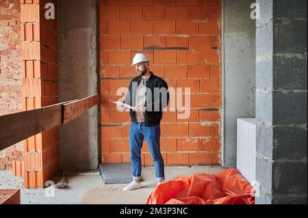 Full length of man worker in safety helmet holding papers and looking away while standing at construction site. Male builder contractor posing against brick wall in building under construction. Stock Photo