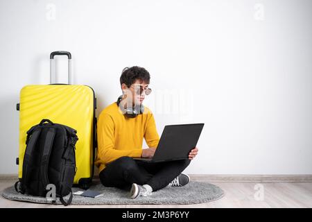 Young guy traveler with a suitcase wears in an orange sweater sits typing on a laptop on isolated white background. freelancer working at the airport. Stock Photo