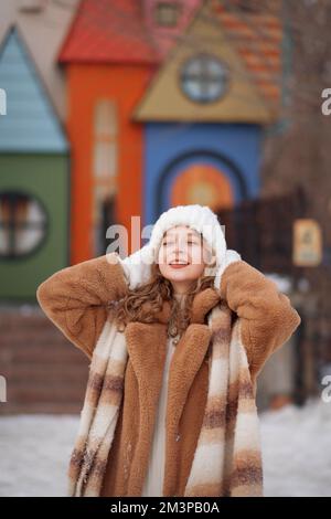 A white blonde girl in a knitted hat, a long scarf and a fur coat walks through the forest and smiles. Woman catches snow in white mittens. Winter is on the street. High quality photo. Christmas Eve in the park. A girl of European appearance enjoys life. New Year's mood, gifts, smiles, sale and joy Stock Photo