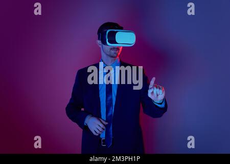 Stylish modern businessman dressed in suit with tie wearing virtual reality glasses on his head standing over gradient background in neon. Stock Photo