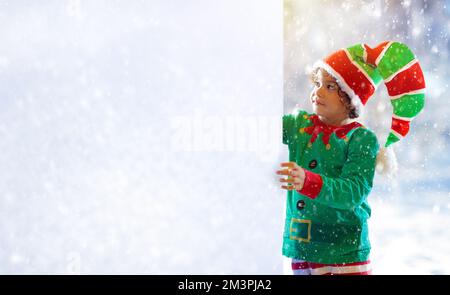 Little boy in elf costume in snowy forest pointing at empty cope space for your text. Merry Christmas card or advertisement template. Gifts for child. Stock Photo