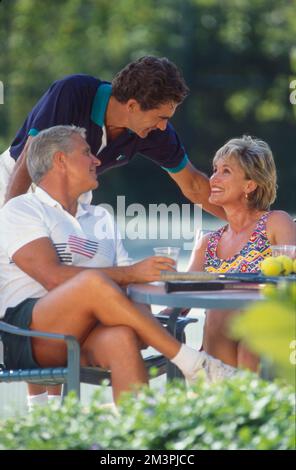 Older couple sitting at table taking a break from tennis game to greet another man Stock Photo