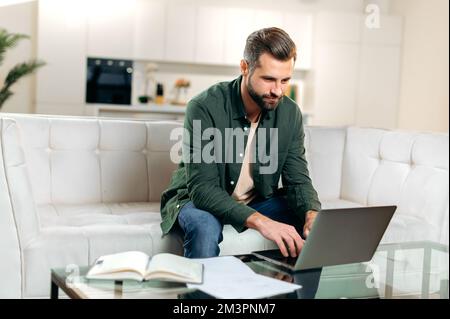 Focused successful caucasian man, freelancer, programmer, sit on a sofa in living room, uses his laptop, working online on a project from home, planning business plan, strategy, searching information Stock Photo