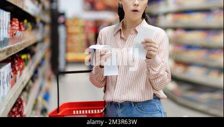 Web banner of shopping and high price. Amazement Caucasian woman holds paper receipts of purchase. In background, rows of shelves with products. Conce Stock Photo