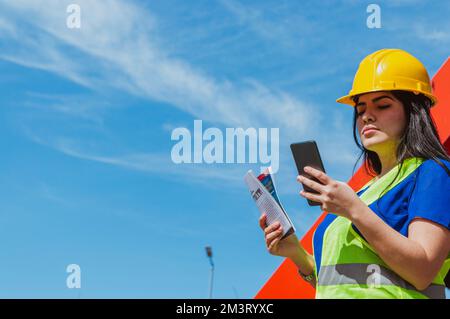 young latin caucasian outdoor worker woman checking notifications on her phone, engineering and industry concept, copy space. Stock Photo