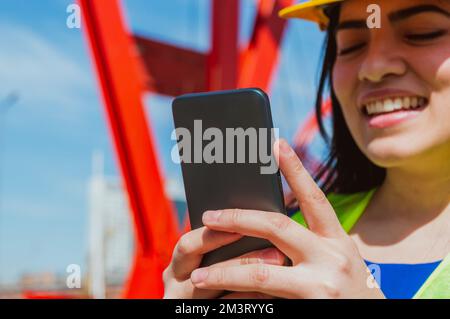 closeup of young female engineer, wearing safety helmet and vest, smiling is outdoors using the phone, focus on the phone. Stock Photo