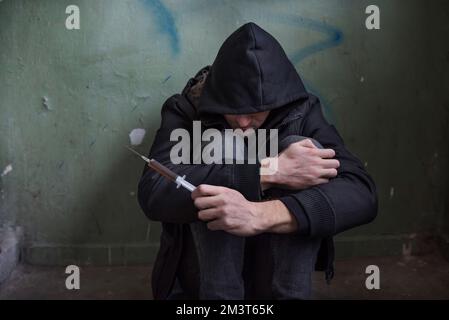 Photo of a desperate young drug addict in the hood and sitting on the floor after using drugs in an abandoned house. Drug abuse concept Stock Photo