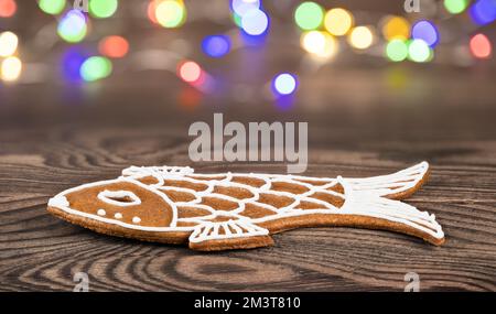 Closeup of Christmas gingerbread cookie in fish shape on wood background. Ornate sweet aromatic pastry for happy Xmas and New Year. LED lights. Bokeh. Stock Photo