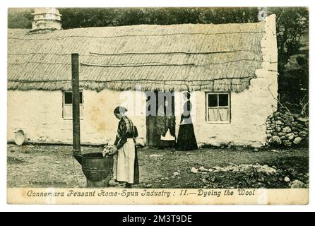 Original Edwardian era greetings postcard of peasant home-spun industry. Dyeing the Wool. outside a rustic cottage home, vernacular architecture built of local stone and roofing of reeds. Connemara, Galway, Republic of Ireland. Circa 1905. Stock Photo