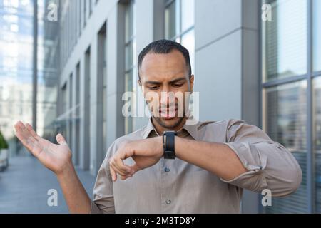 A man is looking unsatisfied at a smartwatch. A Latin American businessman is waiting outside an office building. Stock Photo
