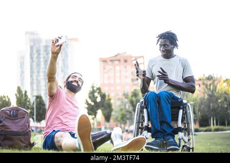 Two young adult friends taking selfie, african man on whellchair and hispanic man with artificial limb having a video call with friends at city park, Stock Photo
