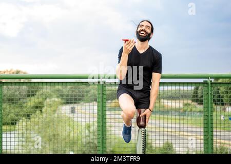 happy man laughing while using phone, young hispanic with an artificial leg relaxing at end workout, person with disability resting after fitness acti Stock Photo