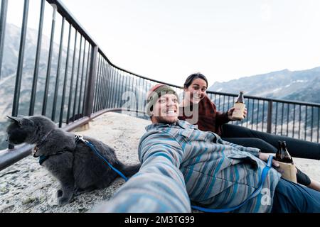 Young 30s caucasian woman sits with her husband and cat enjoying a drink while having her gray russian blue cat on leash in wilderness forest while at Stock Photo
