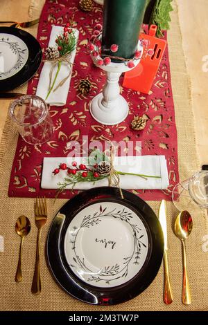 Festive Christmas table setting. On a wooden table lies a red runner, black plates on white says Enjoy, golden forks, spoons, knives. New year green c Stock Photo