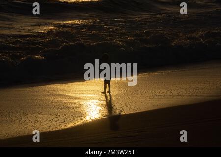 Wanganui New Zealand - April 9 2022; Standing legs of Unrecognizable Young person at waters edge on surf beach in silhouette back-lit by sunset Stock Photo