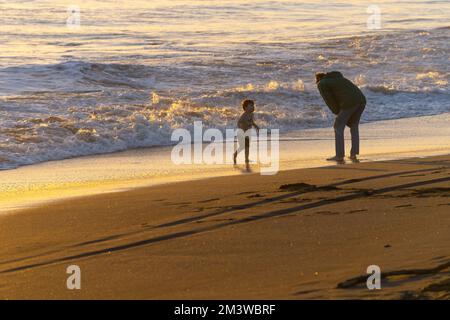 Wanganui New Zealand - April 9 2022; in silhouette father leans down to communicate with son near waters edge Stock Photo