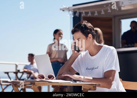 Im waiting for your answer. a cheerful young woman talking on her cellphone while doing work on her laptop next to a beach promenade outside during Stock Photo