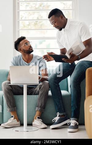I need your help with this. Full length shot of two handsome businessmen sitting together and reading paperwork while using a laptop in the office. Stock Photo