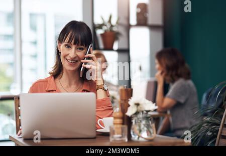 Its so nice to hear from you. an attractive middle aged woman talking on her cellphone while working on a laptop inside of a coffee shop during the Stock Photo