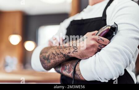 Ill make you look hip and trendy in no time. an unrecognizable tattooed barber posing with his arms folded inside a barbershop. Stock Photo