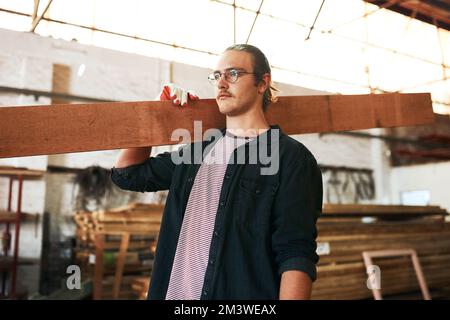 Its all in a days work. a handsome young carpenter carrying a wooden plank over his shoulders inside a workshop. Stock Photo