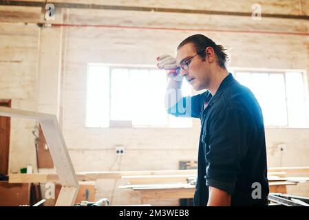 You work up quite a sweat down here. a handsome young carpenter wiping sweat off his forehead while working hard inside a workshop. Stock Photo