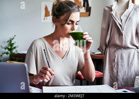 I need all the energy to complete my new collection. a young designer drinking green juice while working at her desk. Stock Photo