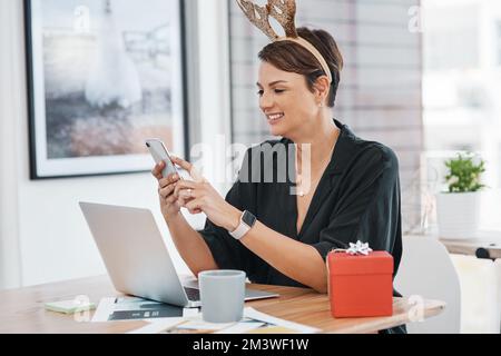 Feeling the Christmas spirit. an attractive young businesswoman sitting and wearing christmas headwear while using her cellphone in the office. Stock Photo