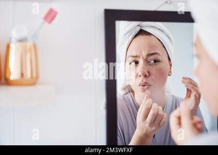 I definitely need to start using moisturizer. a beautiful young woman analyzing her skin in the bathroom mirror. Stock Photo