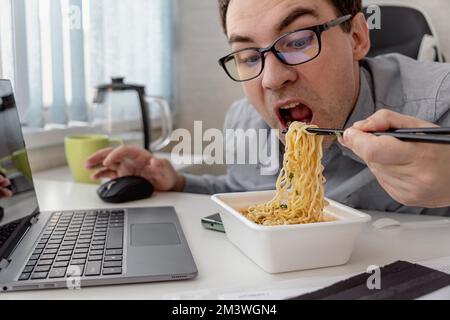 Young man eating instant noodles while working with laptop in office Stock Photo