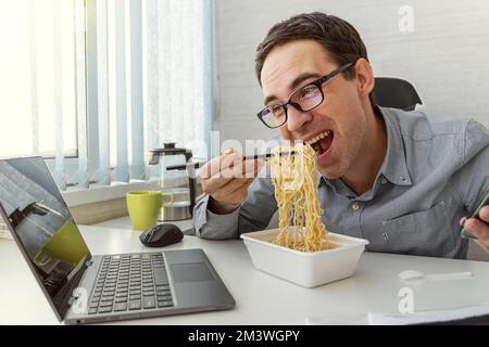 Young man eating instant noodles while working with laptop in office Stock Photo