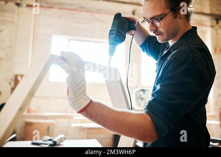 This work of art is almost complete. a handsome young carpenter using an electric drill while working inside a workshop. Stock Photo