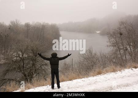 A pensive man in black clothes stands on a high cliff near a river in winter Stock Photo