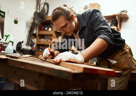 Working hard to earn a living. a focused young carpenter doing measurements on a piece of wood inside of a workshop at night. Stock Photo
