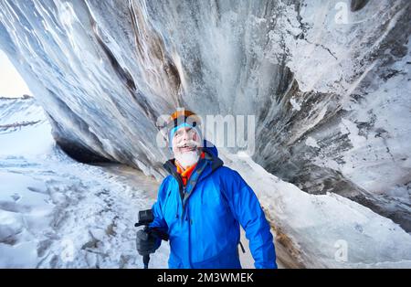 Portrait of smiling old bearded man with action camera in blue costume near ice cave wall at glacier in mountain valley covered with snow in Almaty, K Stock Photo