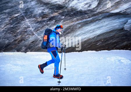 Tourist in blue costume with backpack walking near ice cave textured wall at glacier in beautiful landscape of mountain valley covered with snow in Al Stock Photo