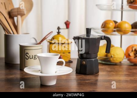Cup of coffee on a kitchen table with a fruit bowl, a jug with wooden utensils, a coffee grinder and an Italian coffee maker. Stock Photo