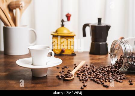 Cup of coffee on a kitchen table with a jug with wooden utensils, a coffee grinder, an Italian coffee maker and an overturned jar with roasted coffee Stock Photo