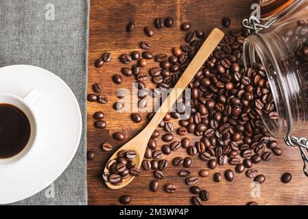 Cup of coffee next to an overturned jar with roasted coffee beans, some on a wooden spoon. Stock Photo