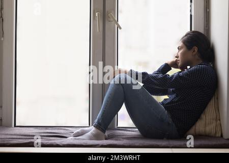 Unhappy lonely young Indian woman sitting on windowsill Stock Photo
