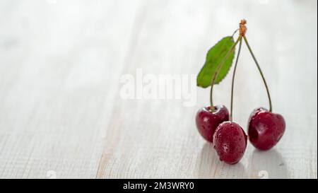 Cherry on white wooden background. Fresh ripe sweet cherry bunch with 3 berries and green leaf with copy space left. Healthy eating, veganism and summer concept. Horizontal banner Stock Photo