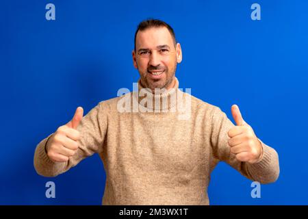 Handsome man wearing brown turtleneck giving thumbs up positive hand gesture smiling and happy for success. Winning gesture Stock Photo