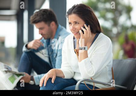 couple waiting at bus-stop reading and using smartphone Stock Photo