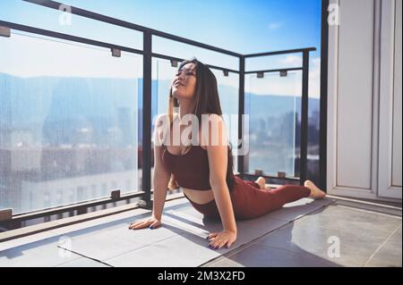 Beautiful Asian smiling fit woman doing yoga on sunny balcony in sport suit. Young happy lady on terrace at summertime. Big city view. Sports exercises and gymnastics, healthy way of life concept Stock Photo